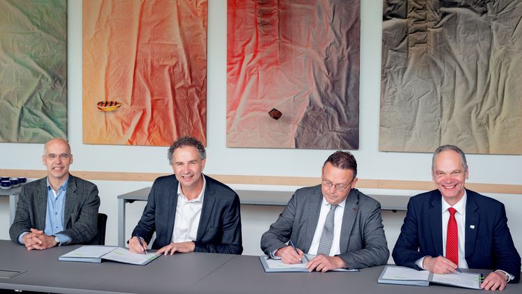 Die vier genannen Personen tragen Anzüge und sitzen nebeneinander an einem Tisch. Jouke de Vries signiert. Im Hintergrund hängen vier bunte Bilder.
